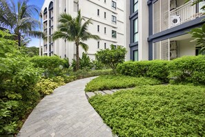 Ein Weg führt zu einem modernen Mehrstöckengebäude, umgeben von üppiger Vegetation und Palmen.