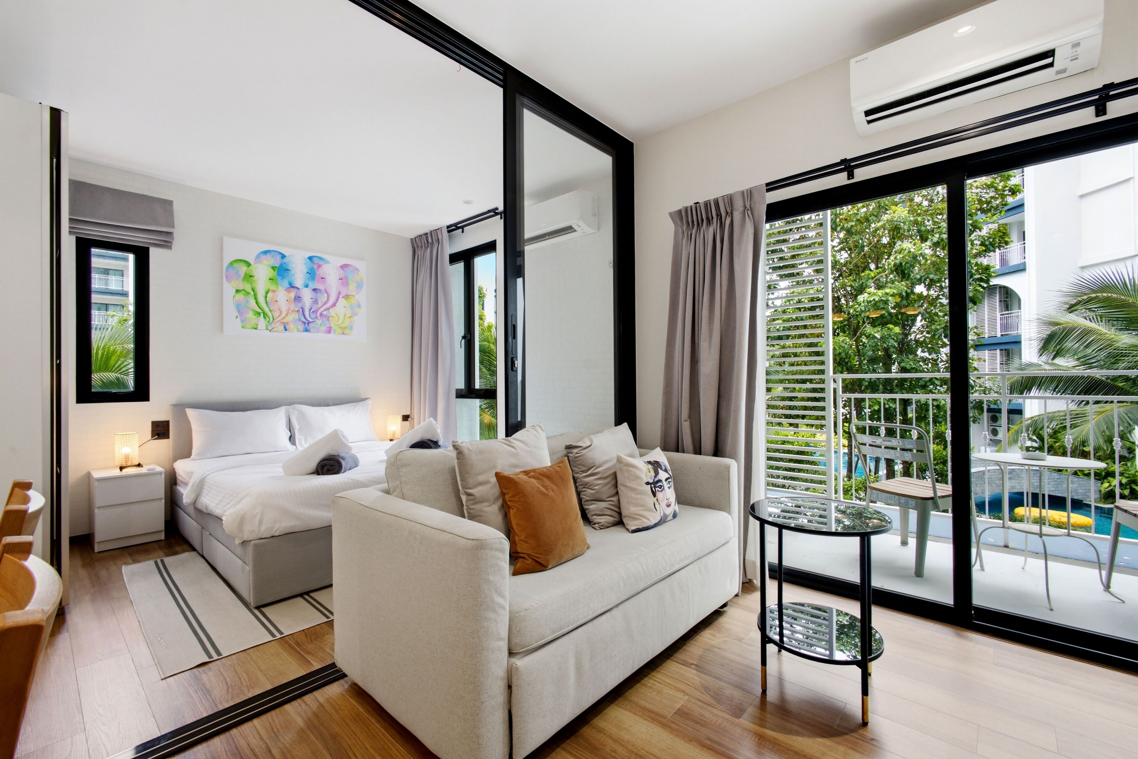 Een moderne hotelkamer met een bed, een bank, een kleine tafel en een balkon met uitzicht op groen en een zwembad.