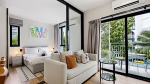 A modern hotel room with a bed, a sofa, a small table, and a balcony with a view of greenery and a pool. - Stylish 1BR Apartment at The Title Halo, Nai Yang (Tambon Sa Khu)