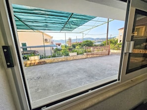 Blick durch ein Fenster mit Metallrahmen, einem Betonboden, einem grünen Metallvordach und einer Steinmauer mit einem Metallgitter.