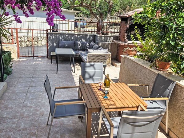 Un patio al aire libre con una mesa y sillas de madera, un sofá con cojines y plantas en macetas.