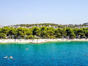 A coastal area with a beach, clear blue water, and a row of buildings with trees in the background. - Lux Adriatica 3 by Interhome (Trogir/Okrug Gornji)