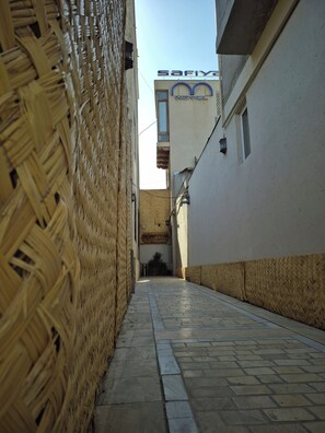 Une ruelle étroite, avec un mur fait de matériaux tressés d’un côté et un bâtiment portant une enseigne indiquant « Hôtel Safiya » de l’autre.