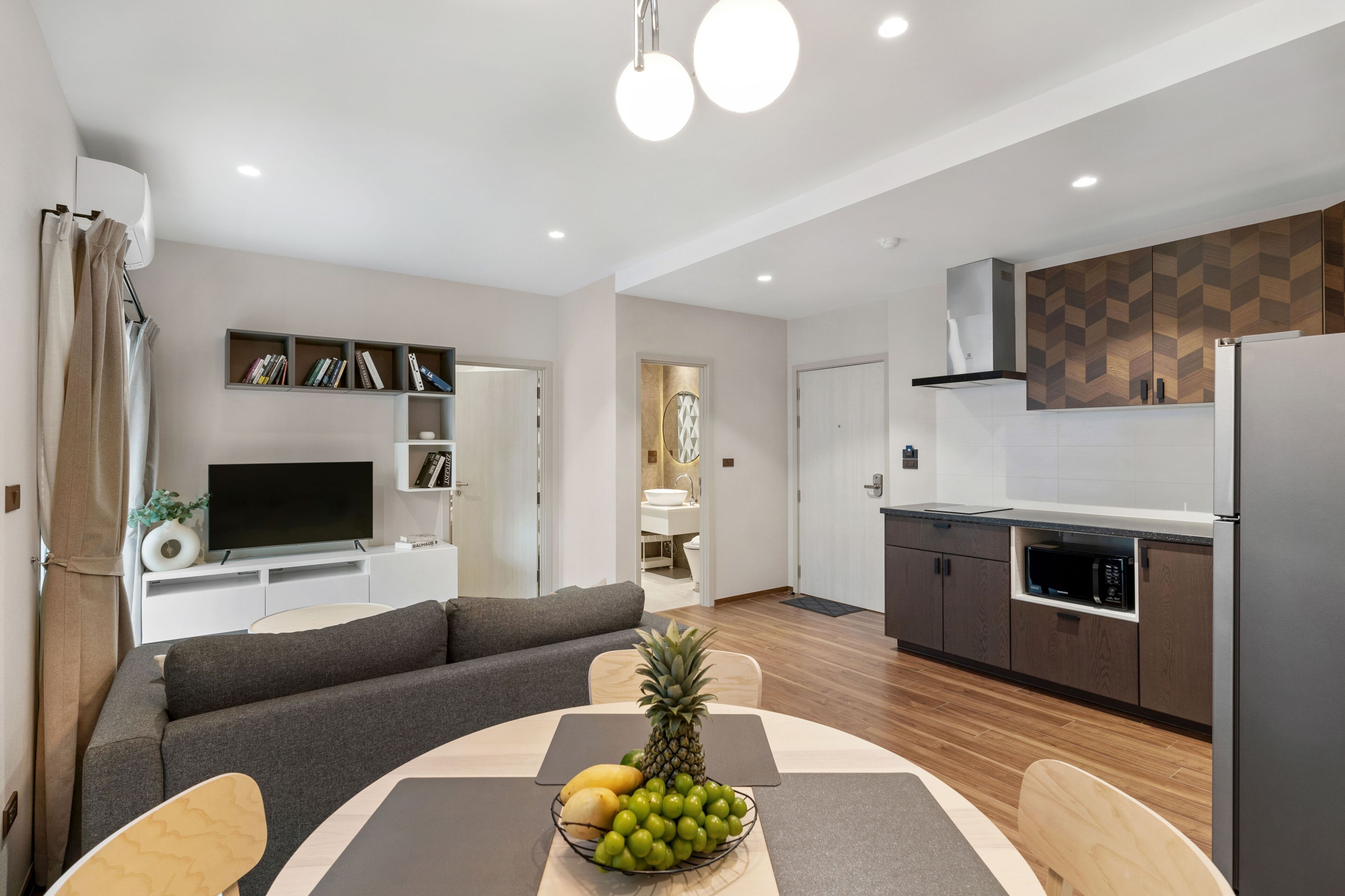 A modern living room with a sofa, a dining table with fruit, a flat-screen TV, and a kitchen with a microwave and refrigerator.