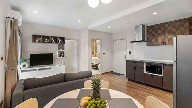 A modern living room with a sofa, a dining table with fruit, a flat-screen TV, and a kitchen with a microwave and refrigerator. - Spacious 2BR at The Title Halo • Walk to Nai Yang Beach (Tambon Sa Khu)