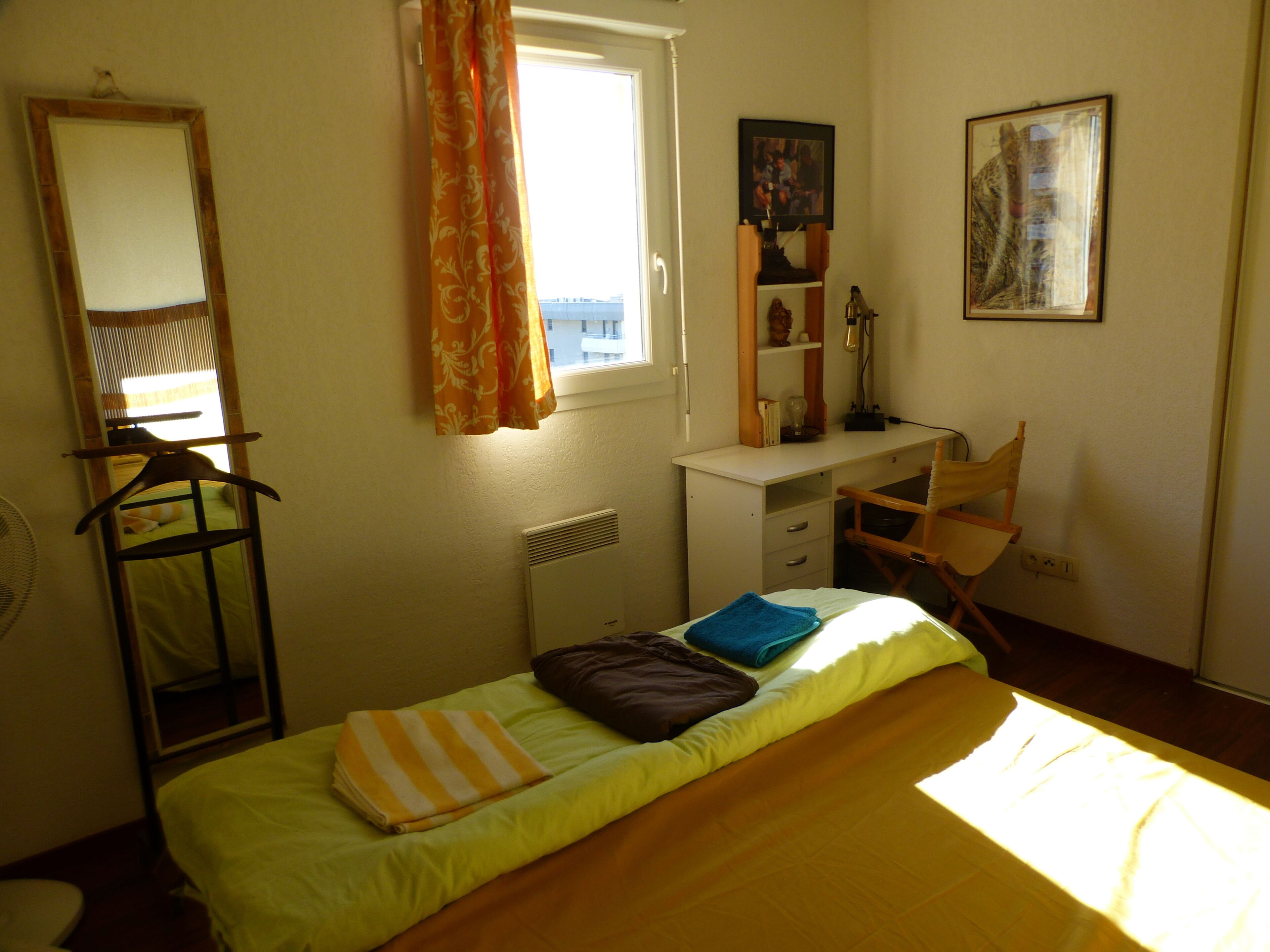 Ein Schlafzimmer mit Bett, Schreibtisch, Stuhl, Heizkörper, einem fenster mit Vorhängen, einem Spiegel und einem Wandregal mit Gegenständen.