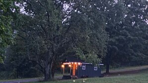 Una cabaña pequeña y oscura con porche y luces, rodeada por un campo cubierto de hierba y árboles.