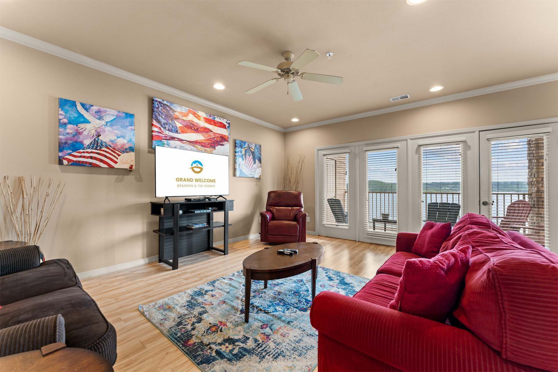 A living room with a red sofa, a brown armchair, a coffee table, and a TV on a stand. There are paintings on the wall and a ceiling fan.