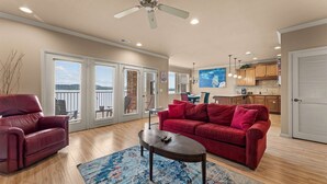 Ein geräumiges Wohnzimmer mit einem roten Sofa, einem Ledersessel und einem Couchtisch. Ein Balkon mit Blick aufs Wasser ist vorhanden, und im Hintergrund ist eine Küchenzeile erkennbar.