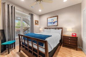 Un dormitorio con cama, mesitas de noche, una silla, ventana con cortinas, ventilador de techo y cuadros en la pared.