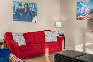 A living room with a red sofa, patterned cushions, a black ottoman, a lamp, and two artworks on the walls. - Yakov's Retreat - Pool Access (Hollister)