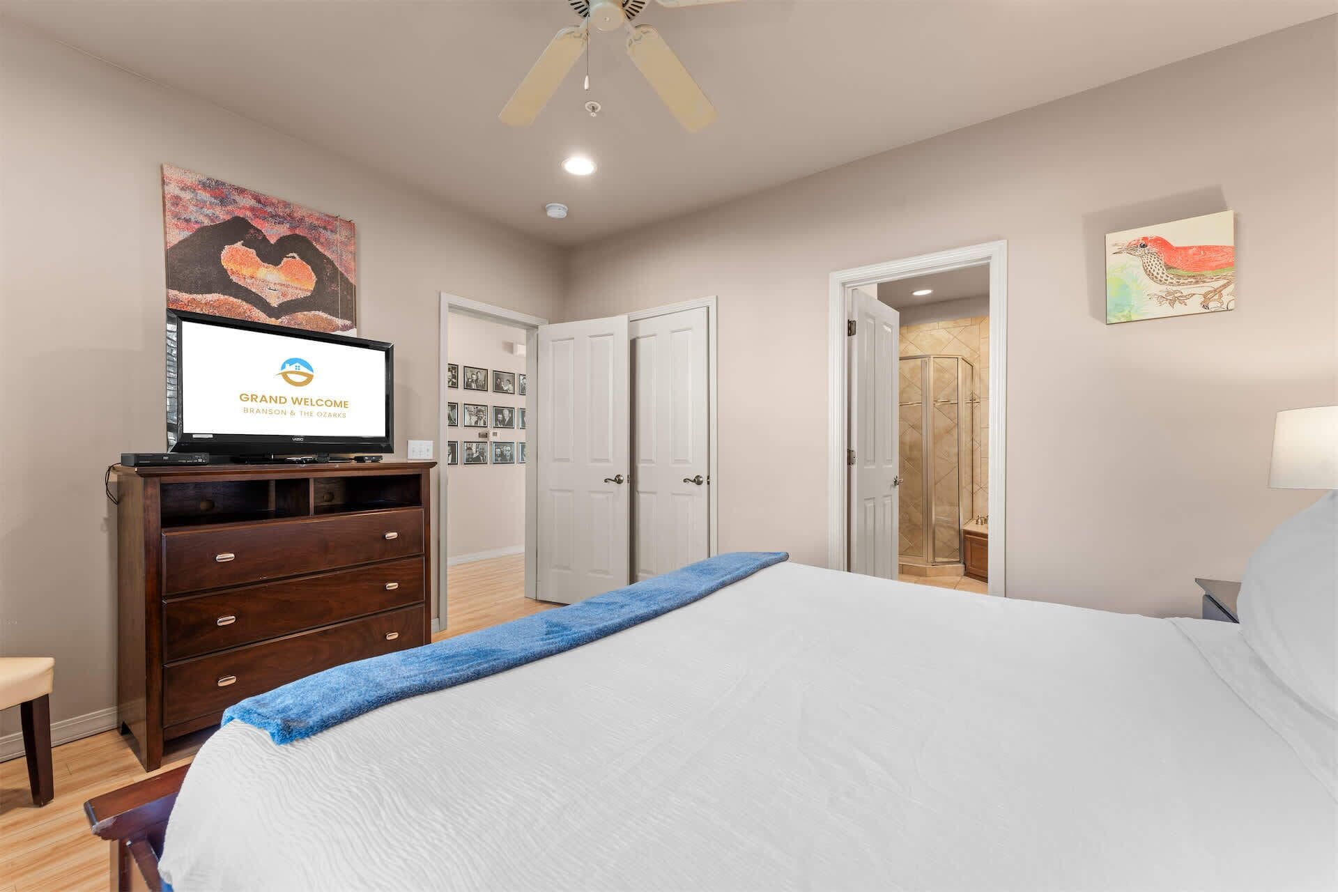 A hotel room with a bed, a television displaying a welcome message, a dresser, a door leading to a bathroom, and a wall with framed pictures.