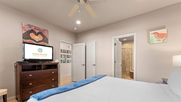 Quarto de hotel com uma cama, uma televisão exibindo uma mensagem de boas-vindas, uma cômoda, uma porta que dá para um banheiro e uma parede com quadros.
