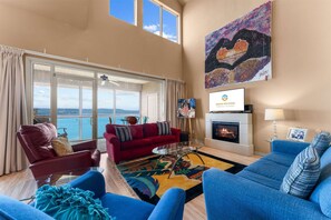 Uma sala de estar espaçosa com uma grande janela com vista para o mar, uma lareira e uma televisão exibindo uma mensagem de boas-vindas.