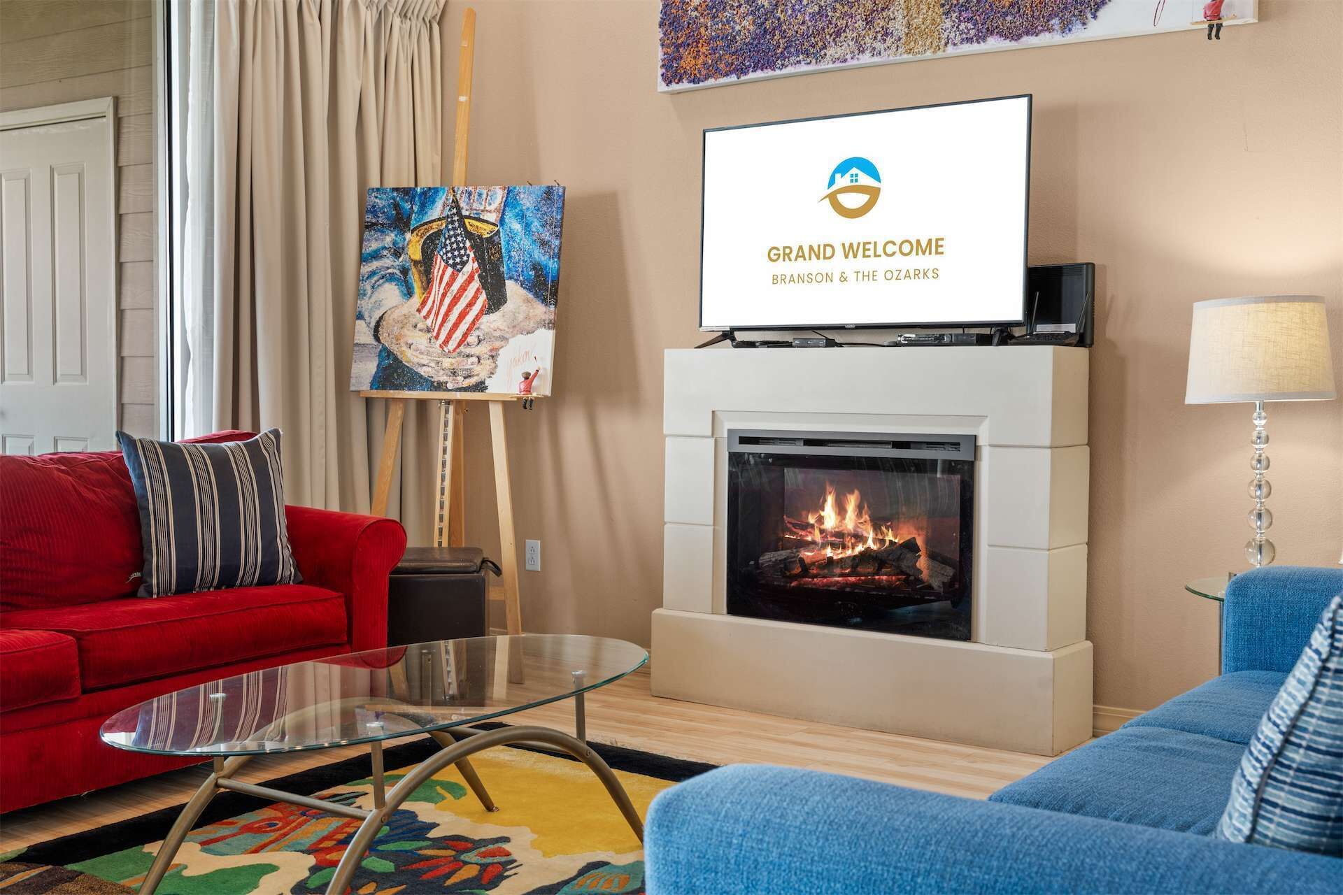 A modern living room with a fireplace, red and blue sofas, a glass coffee table, and a flat-screen TV mounted above the fireplace.