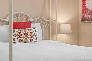 Cama bem arrumada com lençóis brancos e almofadas decorativas, cabeceira de metal branco e luminária de cabeceira.