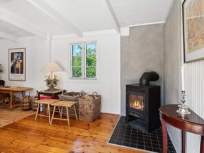 Ein gemütliches Zimmer mit Kamin, Holzboden, Korbmöbeln und einem Fenster mit Grünpflanzen draußen.