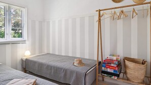 A room with two beds, a window with blinds, a bedside lamp, a hat rack with hats, and a shelf with books and a woven basket. - 5 Star Holiday Home in Graested (Graested)