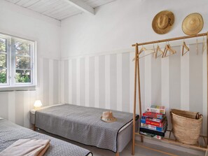 Ein Zimmer mit zwei Betten, einem Fenster mit Jalousien, einer Nachttischlampe, einem Hutfuß mit Hüten und einem Regal mit Büchern und einem gewebten Korb.