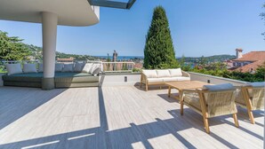Eine Dachterrasse mit einer Couch, einem Tisch und Stühlen. Blick aufs Meer und ein hoher Baum sind zu sehen.