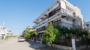 Un edificio residencial de varios pisos con balcones, un coche estacionado en la calle y vegetación a lo largo de la acera.