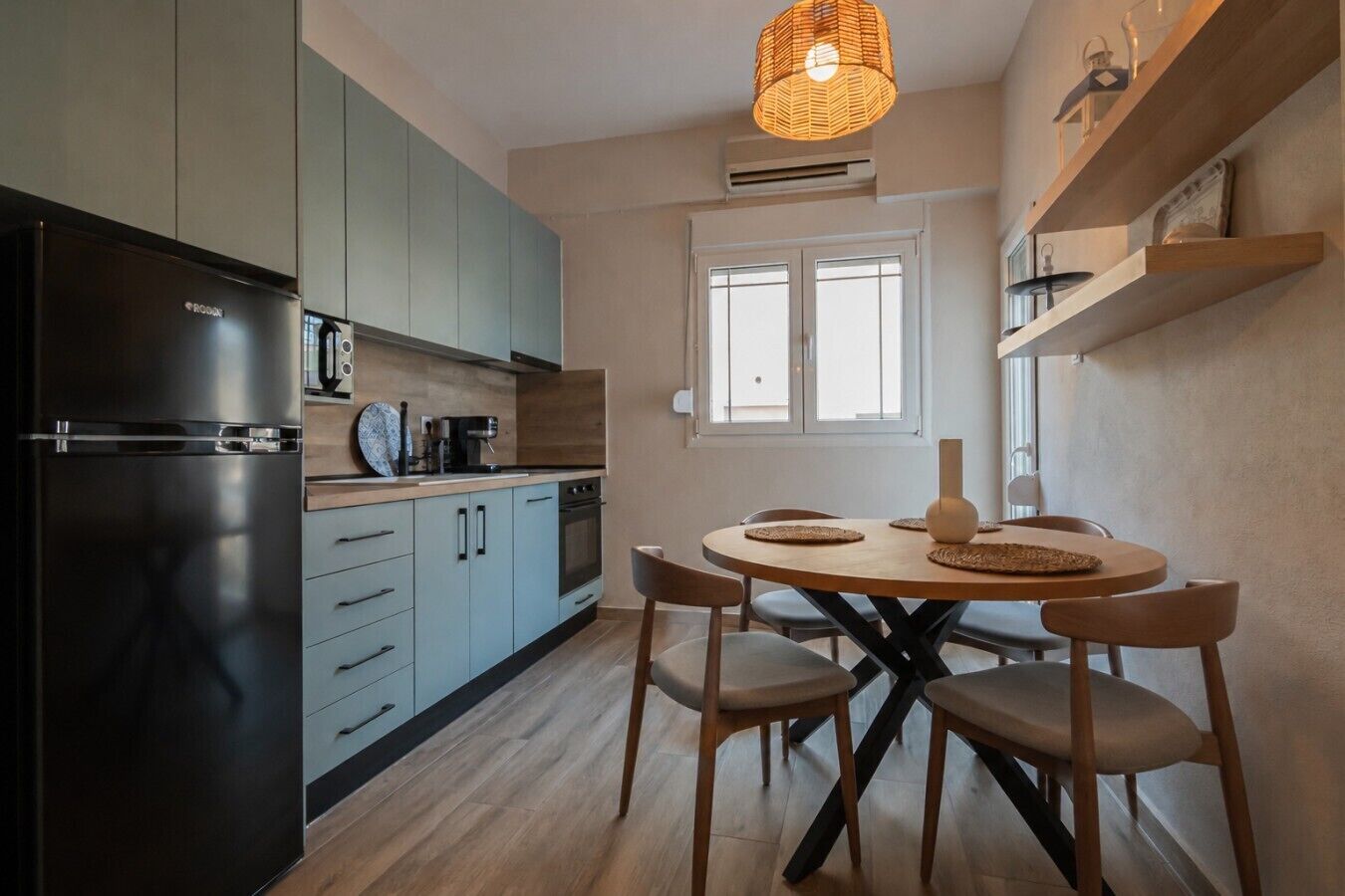 Una cucina moderna con frigorifero nero, mobili azzurri e un tavolo da pranzo in legno con quattro sedie, illuminata da un lampadario.