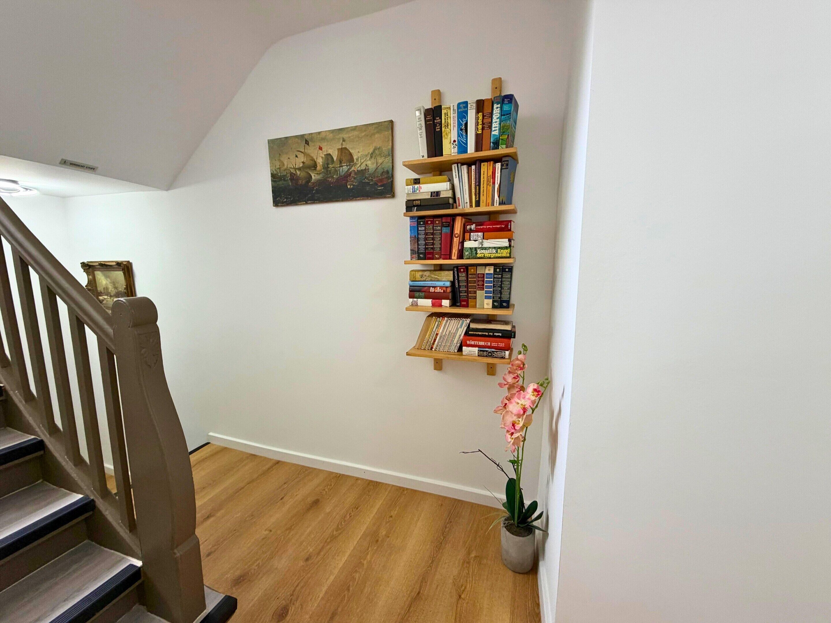 A staircase with wooden steps and a handrail, a bookshelf filled with books, a framed painting on the wall, and a potted plant with pink flowers.