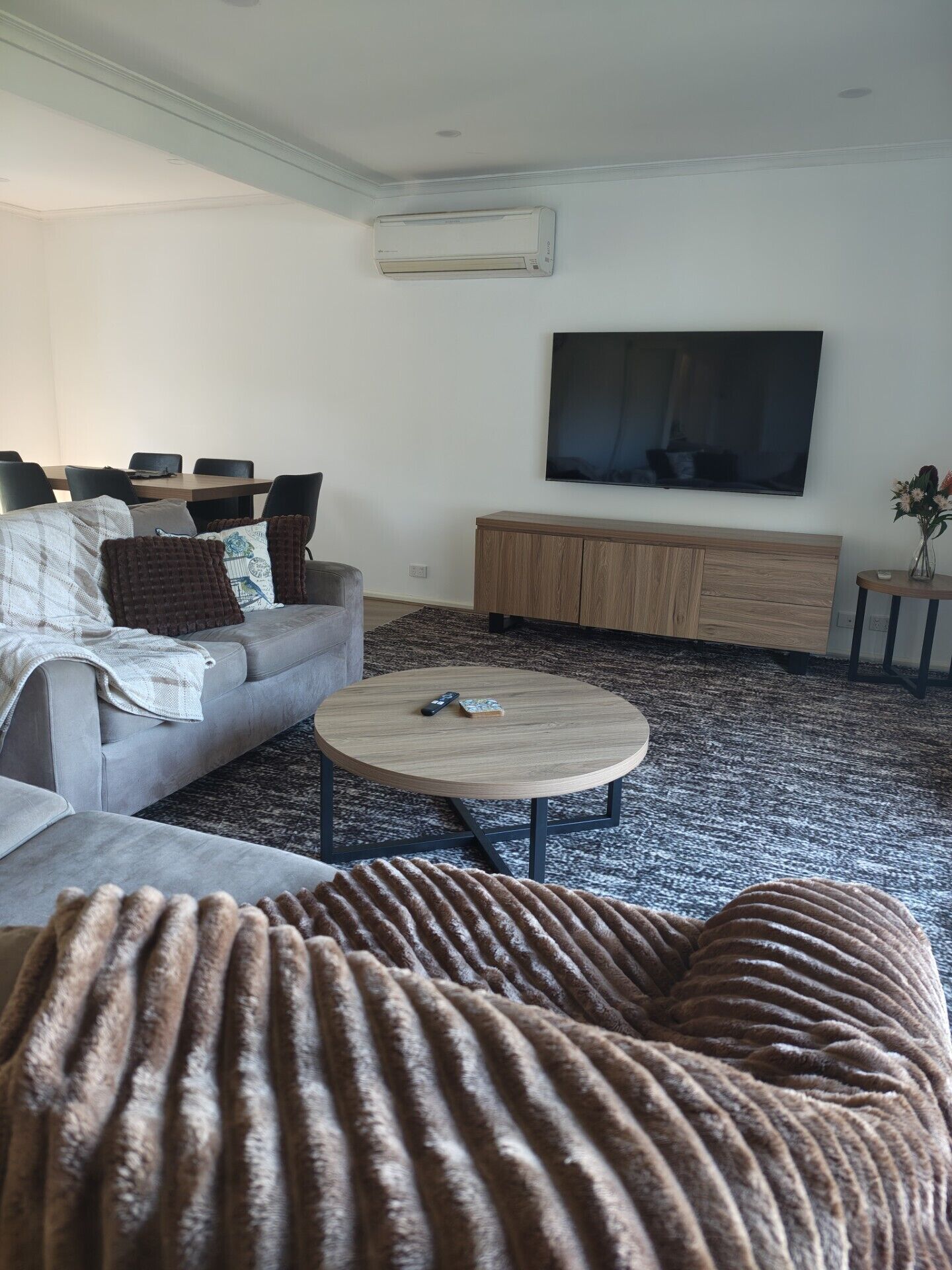 Ein Wohnzimmer mit einer Couch, einem Couchtisch, einem flachbildfernseher an der Wand und einem kleinen Beistelltisch mit einer Vase.