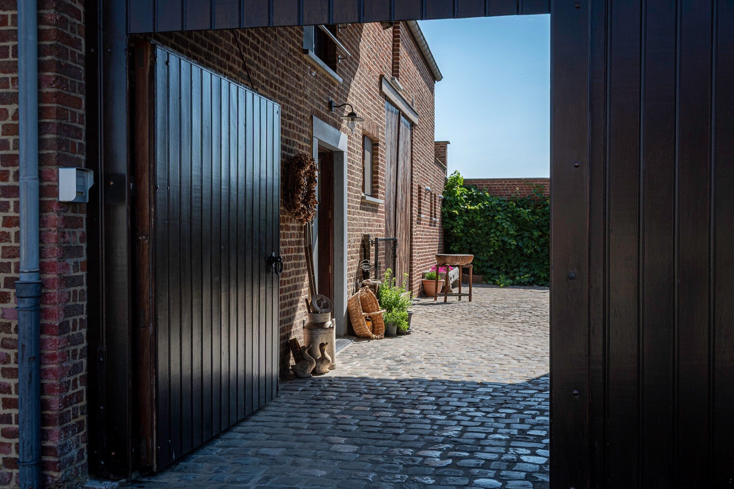 Un edificio de ladrillo con una puerta de madera grande, un pequeño espacio al aire libre con una mesa y sillas, y un camino adoquinado.