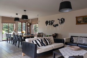 A living room with a sofa, a dining table, and chairs. There is a wall clock and a painting of cows. - Apartment 3 - Enjoy a Cozy Stay at the Alfonsinehoeve (Heers)