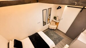Quarto compacto com cama, uma mesinha de cabeceira, espelho e área de chuveiro.