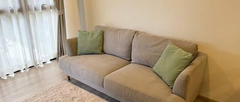 A beige sofa with two green cushions, a beige rug, and a window with sheer curtains.