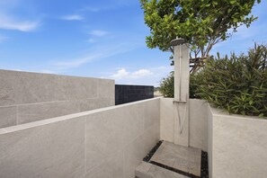 Un área de ducha exterior moderna con un muro de azulejos grises, una sección de azulejos negros y un árbol de hojas verdes.