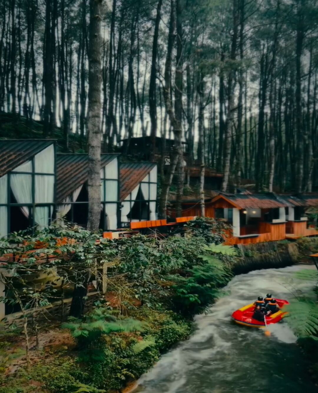 Un río con una balsa, rodeado de bosque denso y edificios con grandes ventanales.