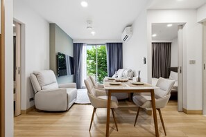 A modern dining area with a wooden table and four chairs, a television mounted on the wall, and a view of greenery outside. - Skypark Laguna 2BR, Golf View, Rooftop Pool (Cherngtalay)