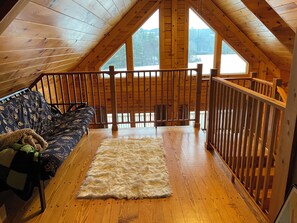 A loft bedroom with a wooden ceiling, a sofa, a fluffy rug, and a view of a snowy landscape. - Cozy 3-bedroom cabin in delightful Monroe with AC and treadmill (Monroe)
