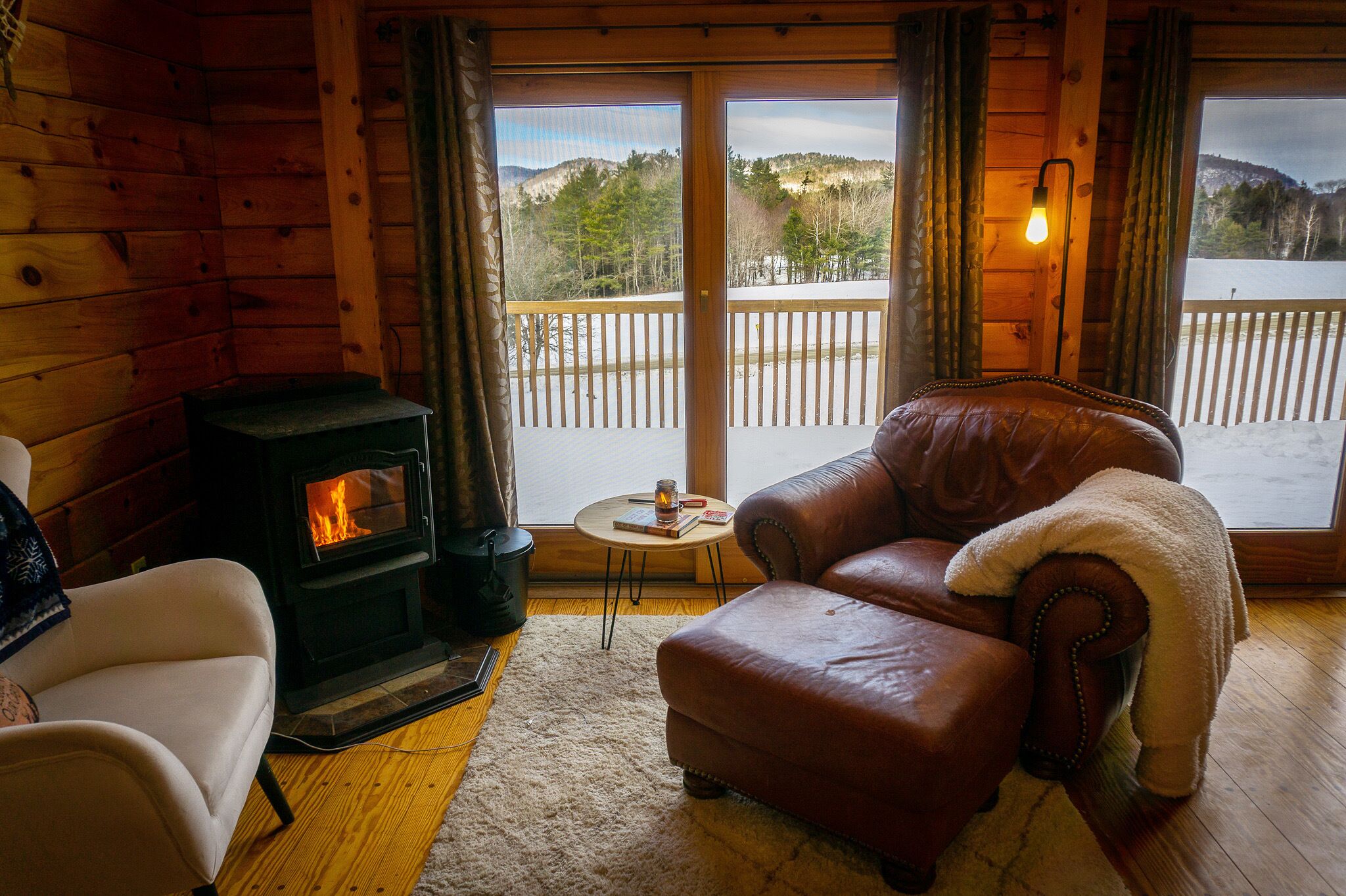L’intérieur chaleureux d’un chalet, avec un poêle à bois, des meubles en cuir et une vue sur un paysage enneigé par de grandes fenêtres.