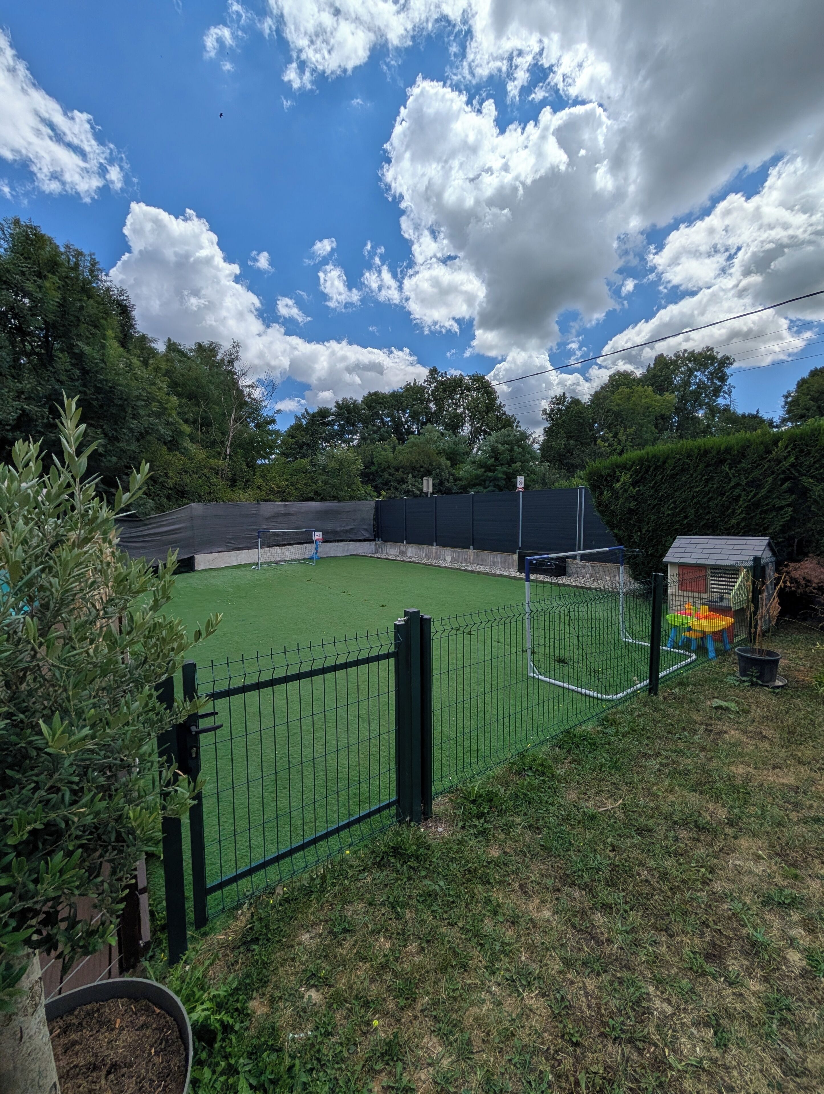 A fenced sports court with a net, surrounded by greenery and trees.