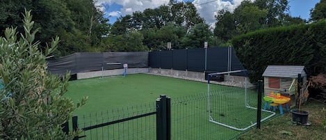 A fenced sports court with a net, surrounded by greenery and trees.