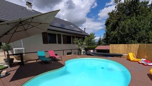 Ein Schwimmbecken mit einer Holzterrasse, einem überdachten Patio und einem Haus im Hintergrund.