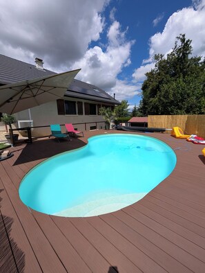 Ein Schwimmbecken mit einer Holzterrasse, einem überdachten Patio und einem Haus im Hintergrund.