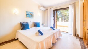 Hotellihuone, jossa on kaksi sänkyä, seinällä taulu, ilmastointilaite ja lasiovesta avautuva näkymä ulos.