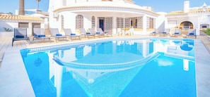 A pool area with lounge chairs and a building with arched windows. - Charming Villa with Heatable Pool and BBQ (Albufeira)