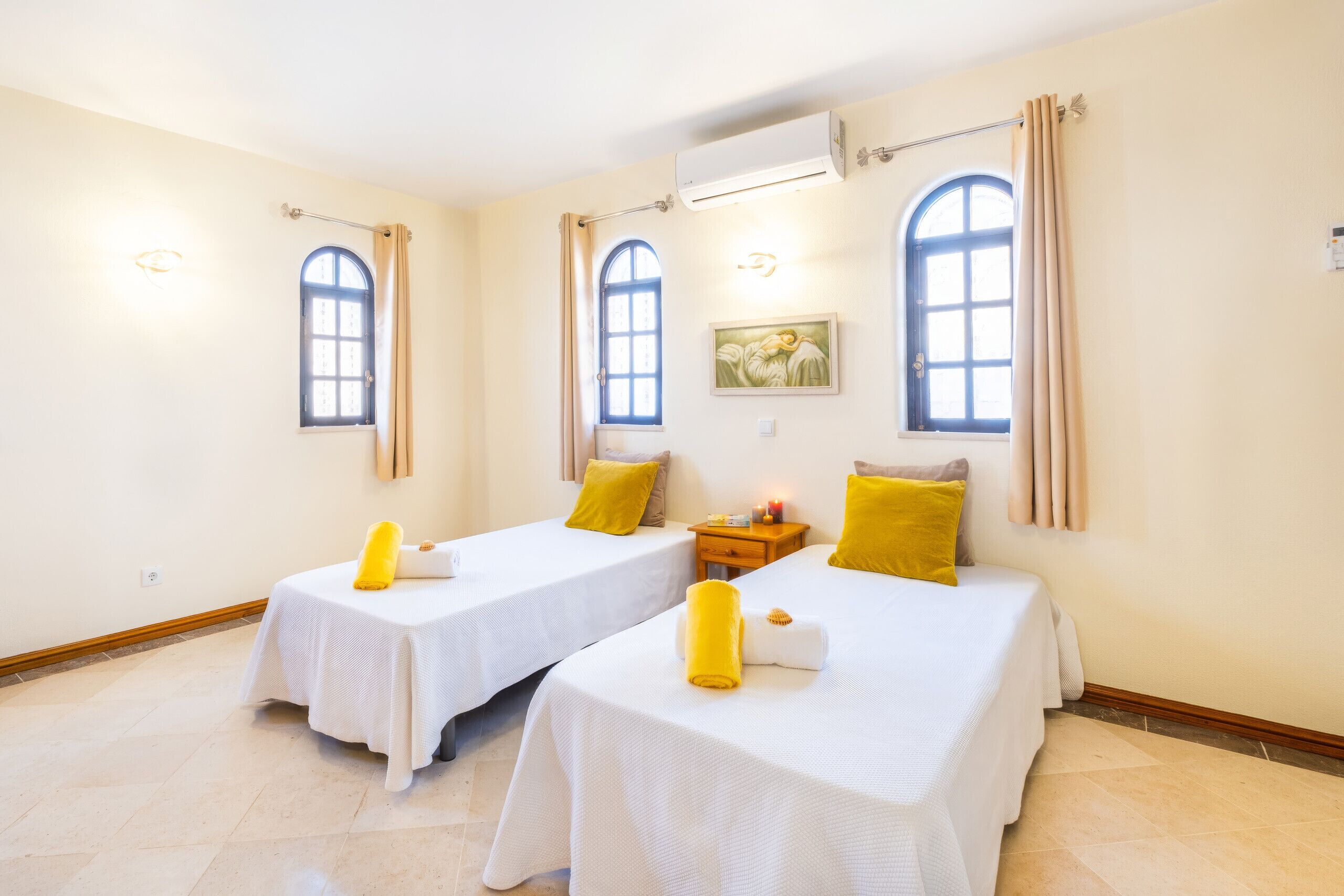 A hotel room with two beds, each with a white bedspread and a yellow pillow. There are two windows with curtains, a wall-mounted air conditioner, and a painting above the beds.