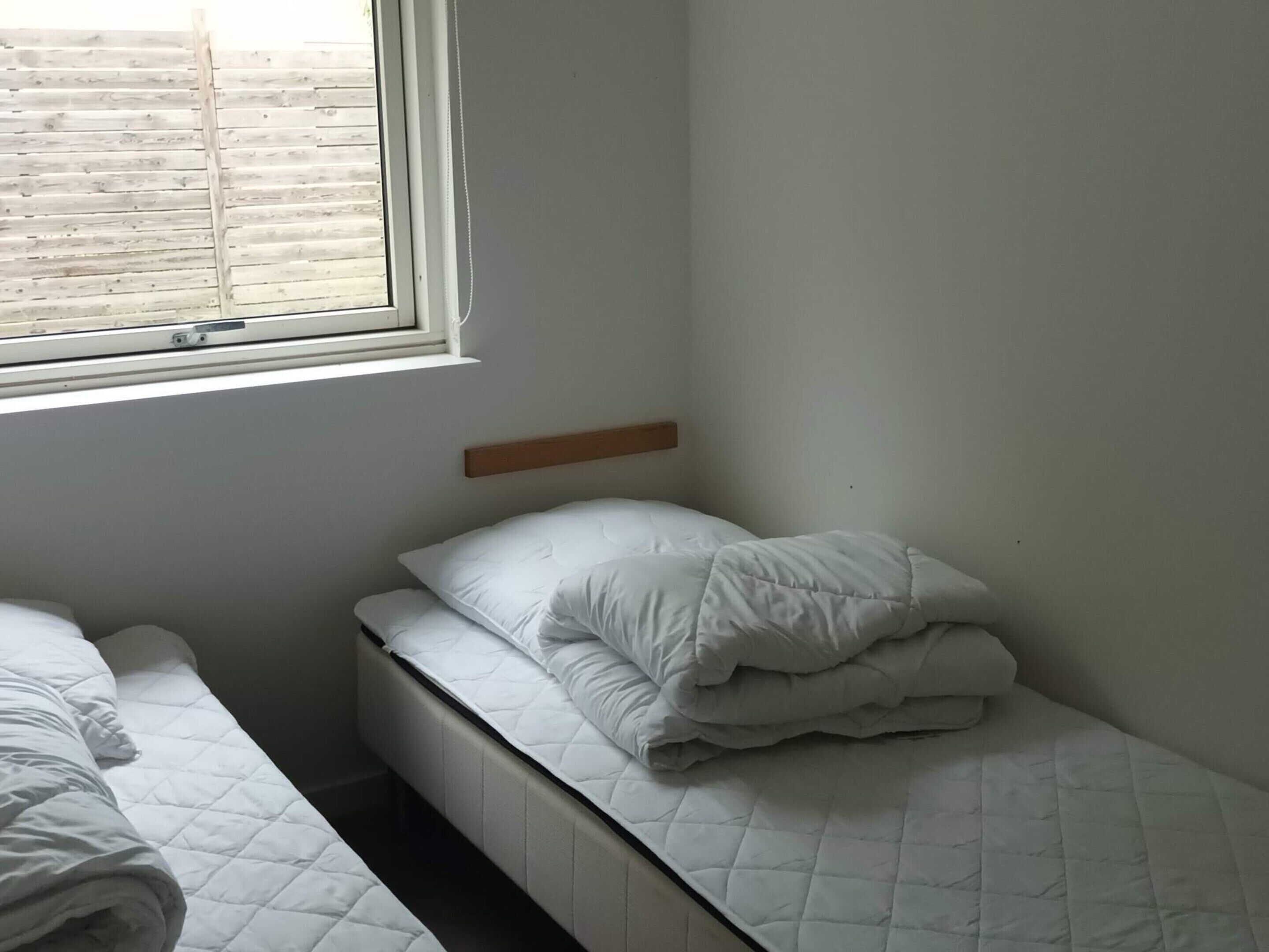 Une petite chambre avec un lit simple, une tête de lit en bois, une fenêtre avec des stores et un mur blanc uni.