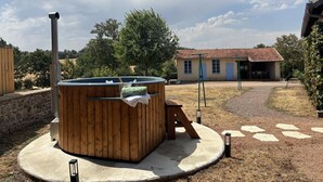 A hot tub in a circular concrete area with a wooden deck, a stone wall, and a house with a tiled roof in the background.