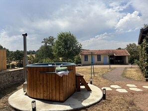A hot tub in a circular concrete area with a wooden deck, a stone wall, and a house with a tiled roof in the background.