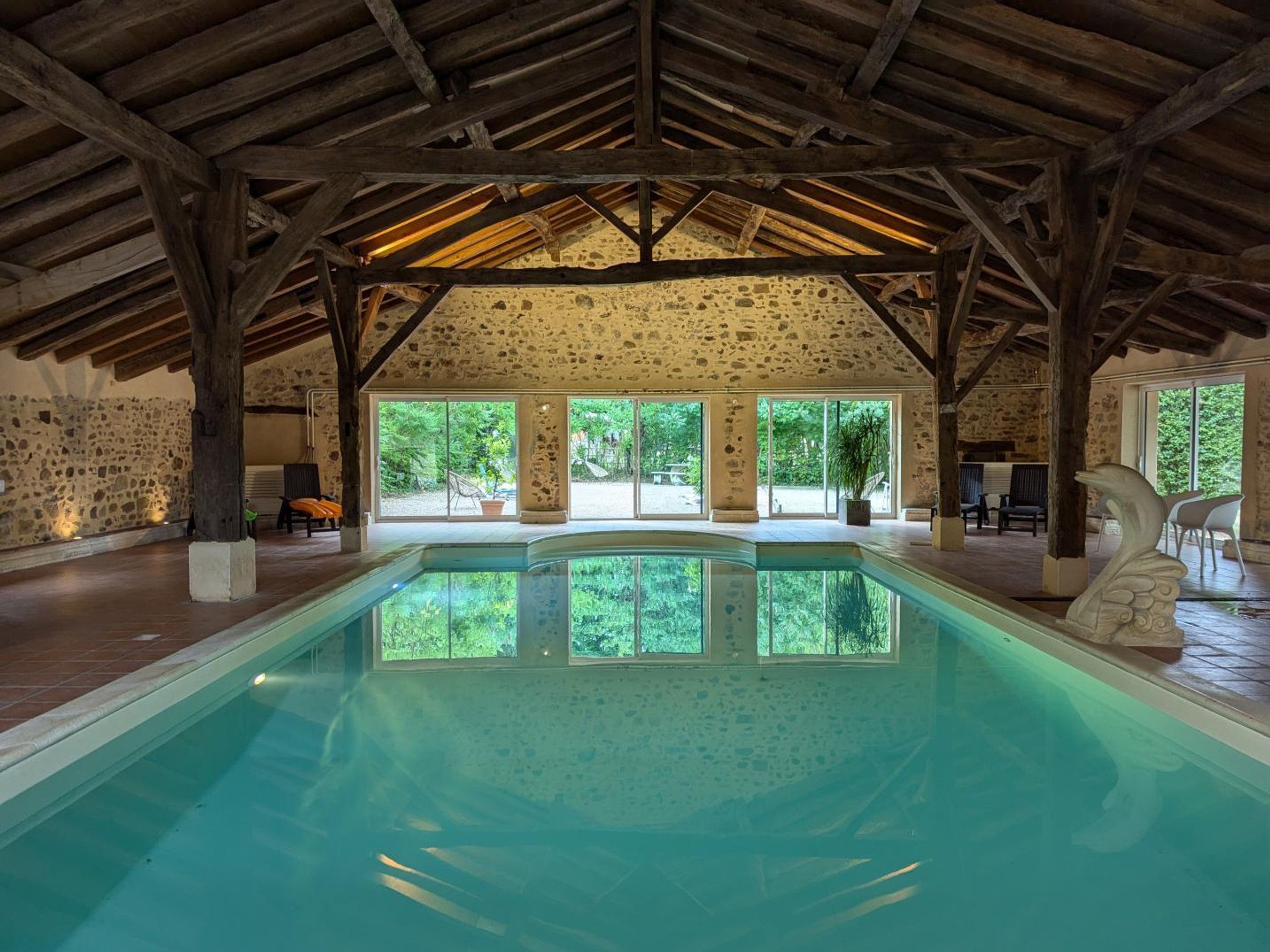 Una piscina con paredes de piedra, vigas de madera, ventanas amplias y mobiliario exterior.