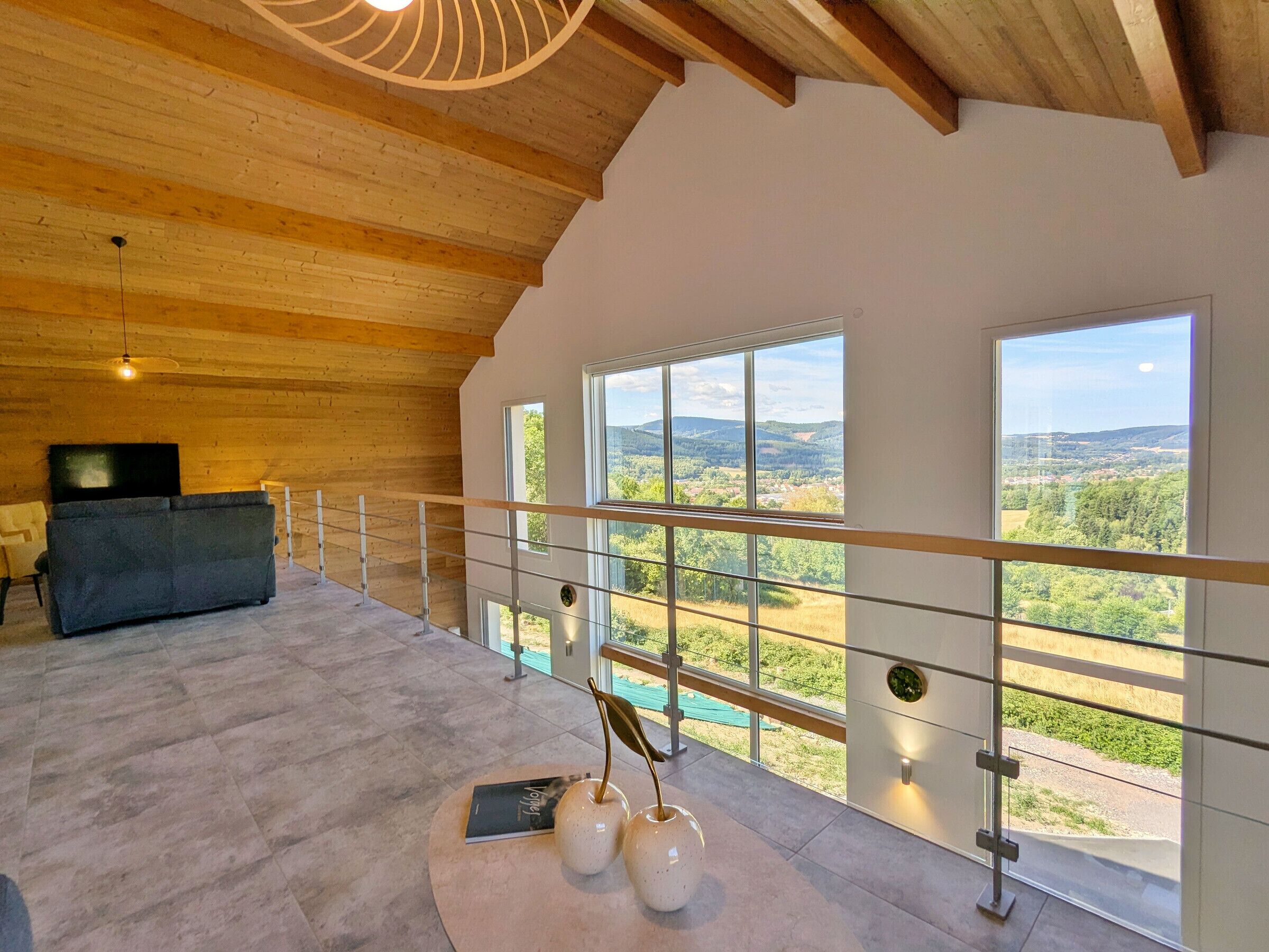 A spacious room with a high ceiling, a sofa, a television, and a view of the countryside through large windows.