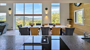 Un coin repas avec une table et des chaises, de grandes fenêtres offrant une vue dégagée et un mur en bois orné d’une horloge.